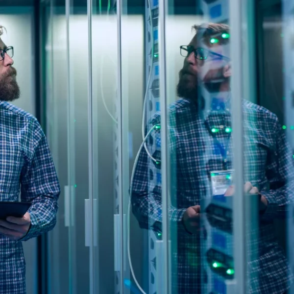 A man holding a tablet while standing in a server room, surrounded by rows of servers and networking equipment.